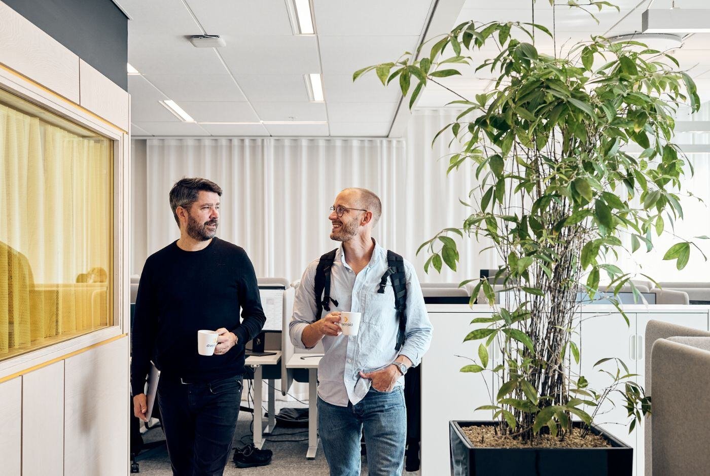 Two male colleagues strolling around the office while holding coffee mugs in their hands